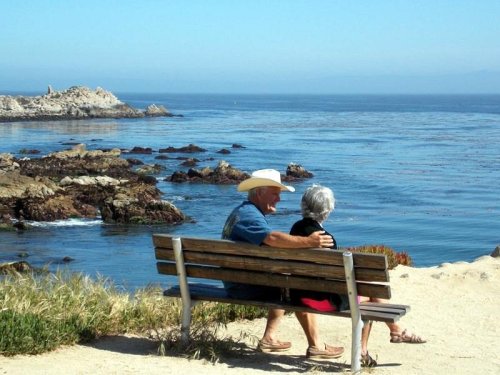 A couple sits on a bench by the ocean, enjoying the view of the water and rocky coastline on a sunny day.
