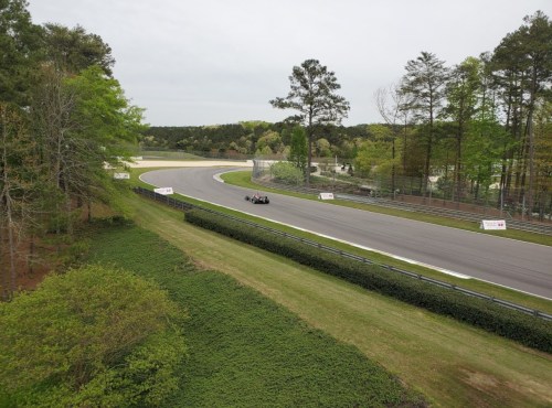 A race car navigates a winding track surrounded by trees and greenery on a cloudy day.
