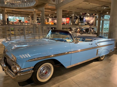 A classic blue convertible car displayed in a modern museum setting, surrounded by motorcycles and automotive exhibits.