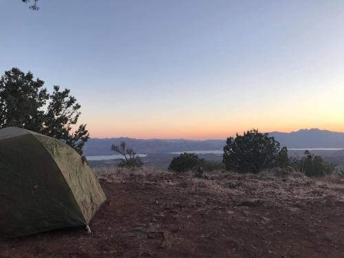 A green tent sits on rocky ground with a scenic view of mountains and a lake at sunset.