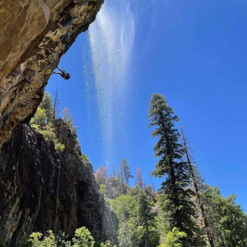 A climber ascends a rocky cliff with a waterfall cascading nearby, surrounded by tall trees and a clear blue sky.