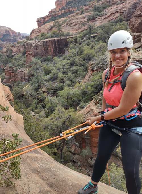 A woman in climbing gear rappels down a rocky cliff with a lush canyon in the background.