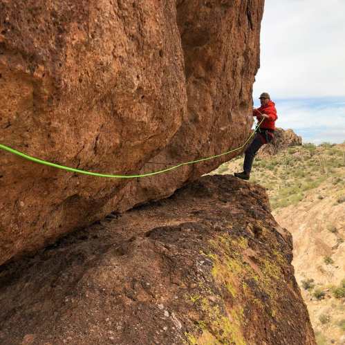 A climber in a red jacket ascends a rocky cliff, secured by a green rope against a backdrop of desert landscape.