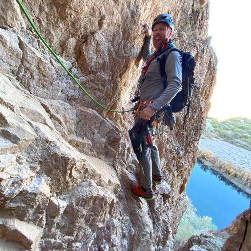 A climber ascends a rocky wall, wearing a helmet and harness, with a scenic view of water and vegetation below.