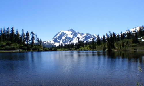 A serene lake reflects a snow-capped mountain, surrounded by lush green trees under a clear blue sky.