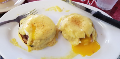 Two eggs Benedict on a white plate, topped with creamy hollandaise sauce and a runny yolk, served with a fork.