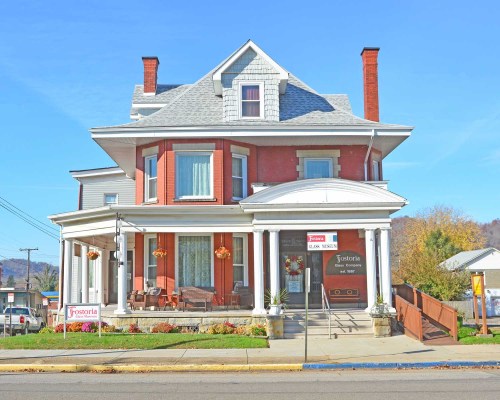 A large, historic red brick house with a gray roof, featuring a porch and flower baskets, set against a clear blue sky.