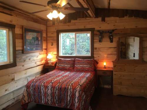 Cozy bedroom with wooden walls, a patterned bedspread, windows, and rustic furniture, illuminated by soft lighting.