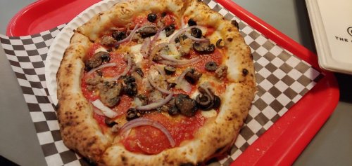 A freshly baked pizza topped with pepperoni, olives, onions, and cheese on a checkered paper plate.