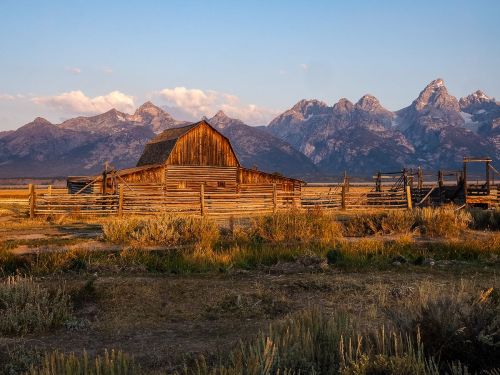A rustic wooden barn surrounded by a fence, with majestic mountains in the background under a colorful sky.