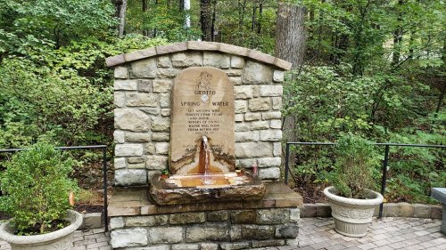 A stone fountain with a plaque reading "Spring Water" surrounded by greenery and potted plants.