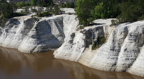 A steep, white cliffside reflected in brown water, surrounded by greenery and a clear blue sky.
