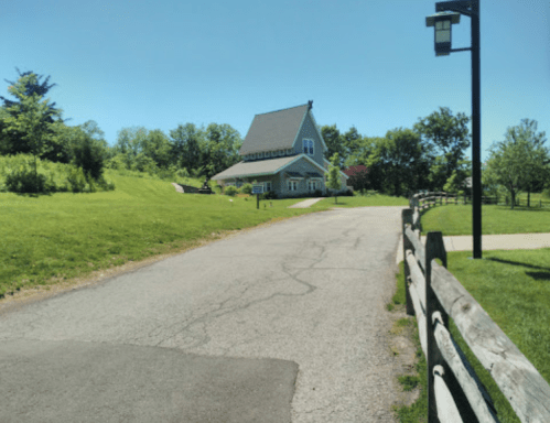 A scenic view of a house on a grassy hill, with a paved road and trees under a clear blue sky.