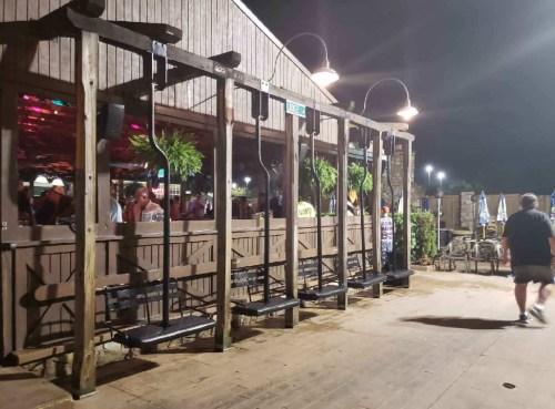 Outdoor seating area with swings, wooden structure, and colorful lights at night. People are visible inside the venue.