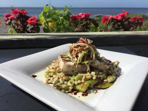 A plate of grilled fish atop couscous and vegetables, with a scenic ocean view and colorful flowers in the background.