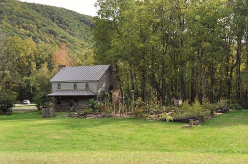 A rustic two-story house surrounded by trees and a garden, set against a backdrop of rolling green hills.