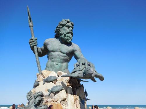 A large bronze statue of a muscular figure holding a trident, surrounded by sea creatures, against a clear blue sky.