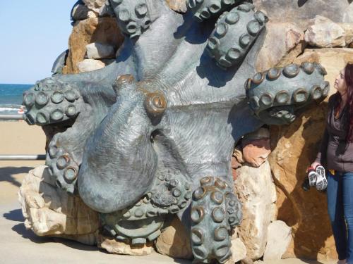 A large bronze octopus sculpture on a rocky wall, with a person standing nearby, against a beach backdrop.