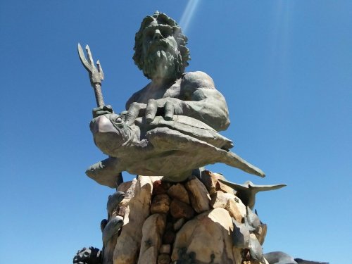 A large statue of a muscular figure holding a trident, seated on rocks with a turtle beneath him, against a clear blue sky.