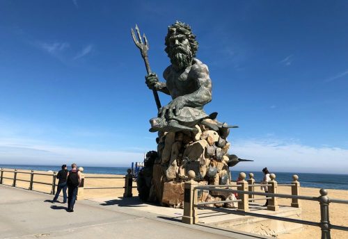 A large bronze statue of Neptune holding a trident, overlooking a sandy beach and ocean under a clear blue sky.