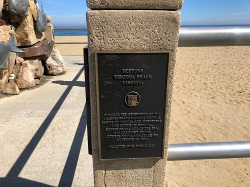 Plaque at Neptune, Virginia Beach, honoring the festival and its supporters, with a sandy beach and ocean in the background.
