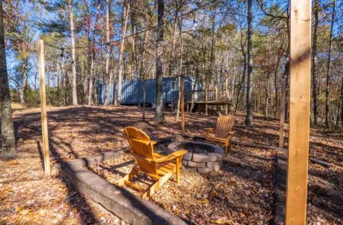 Two wooden chairs around a stone fire pit in a wooded area with a cabin in the background. Autumn leaves scattered on the ground.