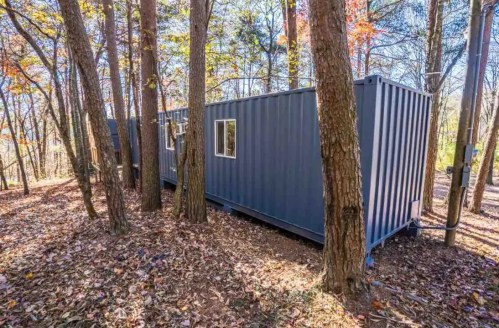 A blue shipping container home nestled among trees in a wooded area, surrounded by fallen leaves.