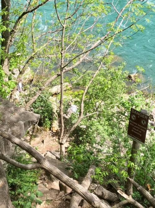 A rocky area near a river, surrounded by trees and greenery, with a warning sign about danger nearby.