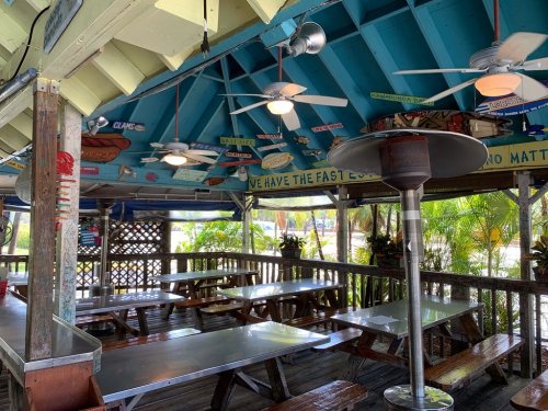 A colorful outdoor dining area with wooden tables, ceiling fans, and tropical decor under a bright blue roof.