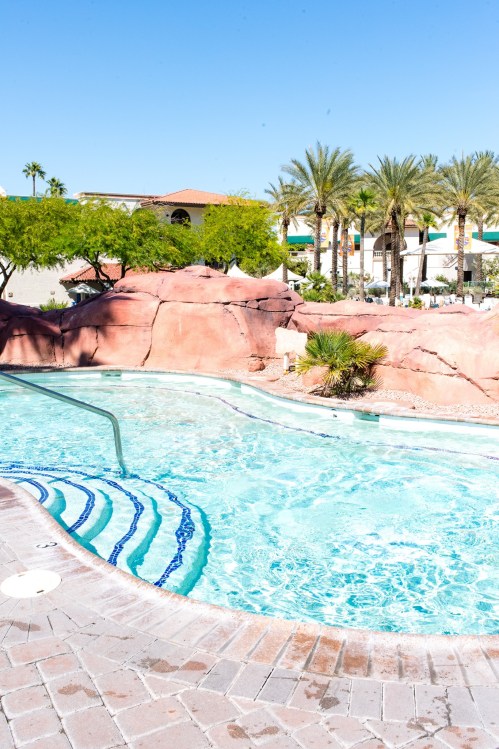 A sparkling pool surrounded by palm trees and rocky landscaping under a clear blue sky.