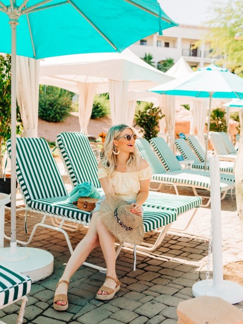 A woman in a sun hat and sunglasses sits by a poolside, surrounded by striped lounge chairs and umbrellas.