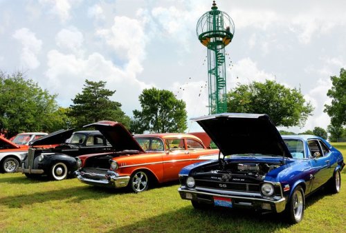 A lineup of classic cars with hoods open, set against a green landscape and a tall, vintage-style tower.