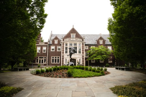 A grand brick mansion surrounded by lush greenery and a circular flower bed in the foreground.