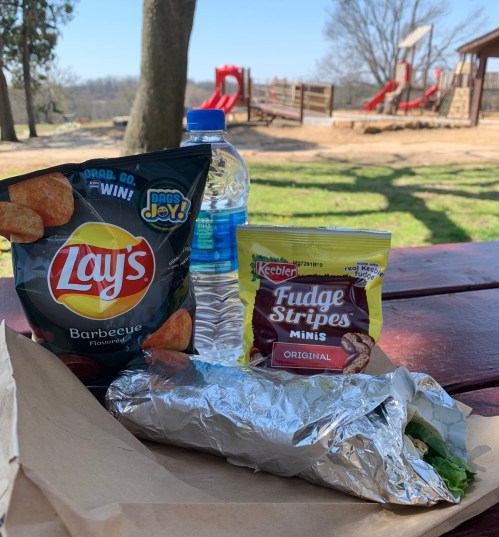 A picnic table with a bag of Lay's barbecue chips, Keebler Fudge Stripes, a water bottle, and a wrapped sandwich.