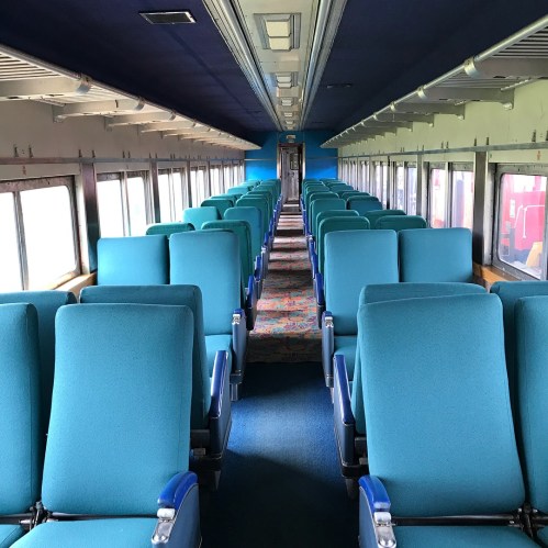Interior of a train car featuring rows of teal seats and a patterned carpet, with large windows on one side.