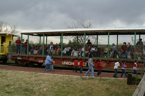A group of people on a train car, with children walking nearby, set against a cloudy sky.