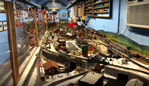 A model train layout with buildings, tracks, and scenery, featuring people interacting in a colorful room.