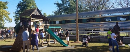 A playground with a slide and climbing structure, next to a train and people enjoying the park on a sunny day.