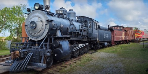 A vintage steam locomotive numbered 643, parked on tracks with a red caboose in the background under a cloudy sky.