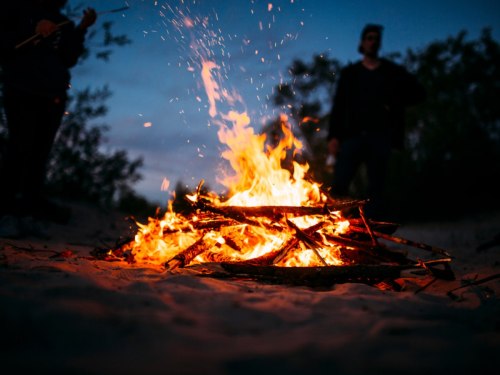A vibrant campfire burns brightly at dusk, with silhouettes of people in the background.