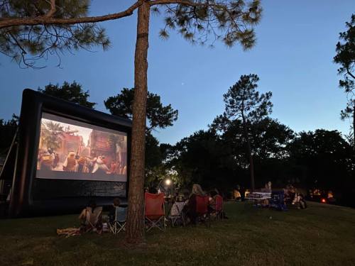 A group of people watching a movie on an inflatable screen in a park at dusk, surrounded by trees.