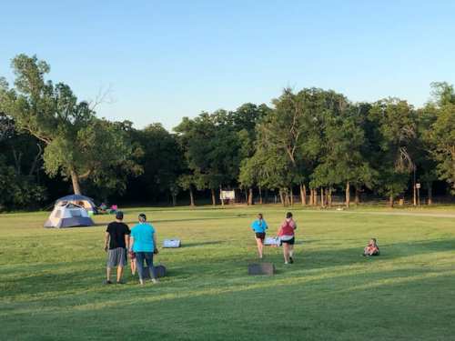 A group of people set up a tent in a grassy area, surrounded by trees, with some engaging in activities nearby.