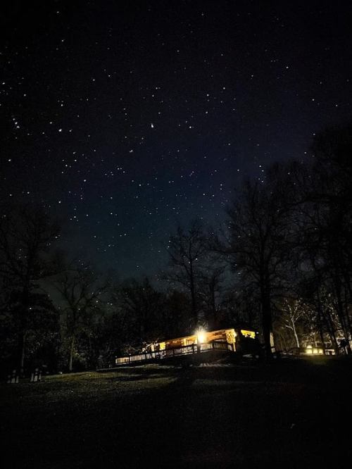 A cabin illuminated at night under a starry sky, surrounded by dark trees.