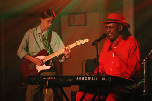 A musician in a red outfit plays keyboard while a guitarist in a green shirt performs on stage. Colorful lights illuminate the scene.