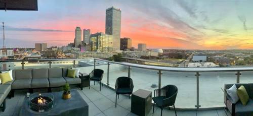 Rooftop view of a city skyline at sunset, featuring modern buildings and a cozy seating area with a fire pit.