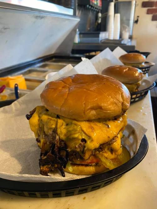 A towering cheeseburger with melted cheese, crispy toppings, and a soft bun, served in a basket with parchment paper.