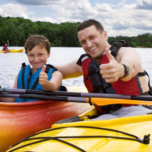 A man and a boy in colorful kayaks give thumbs up, smiling happily on a lake with trees in the background.