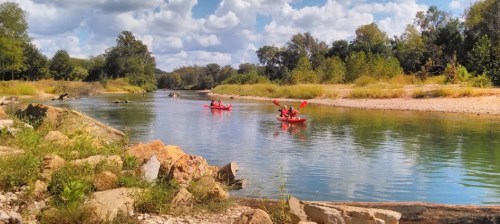 Two kayakers in red kayaks paddle along a calm river surrounded by lush greenery and a blue sky with fluffy clouds.