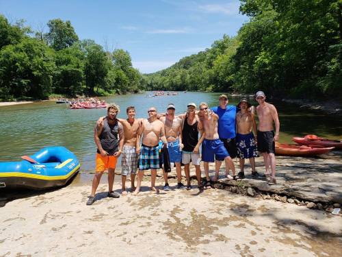 A group of men in swimwear stands by a river, with kayaks and rafts in the background on a sunny day.