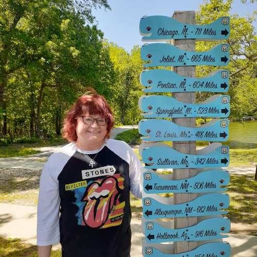 A woman stands next to a signpost with directional arrows and distances to various cities along Route 66.